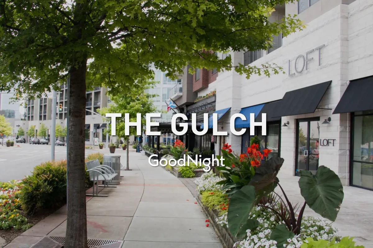 A tree-lined sidewalk leads past colorful flower beds and inviting storefronts, with benches for relaxing. The atmosphere is welcoming and encourages strolling, shopping, and discovering hidden gems in a vibrant neighborhood setting.