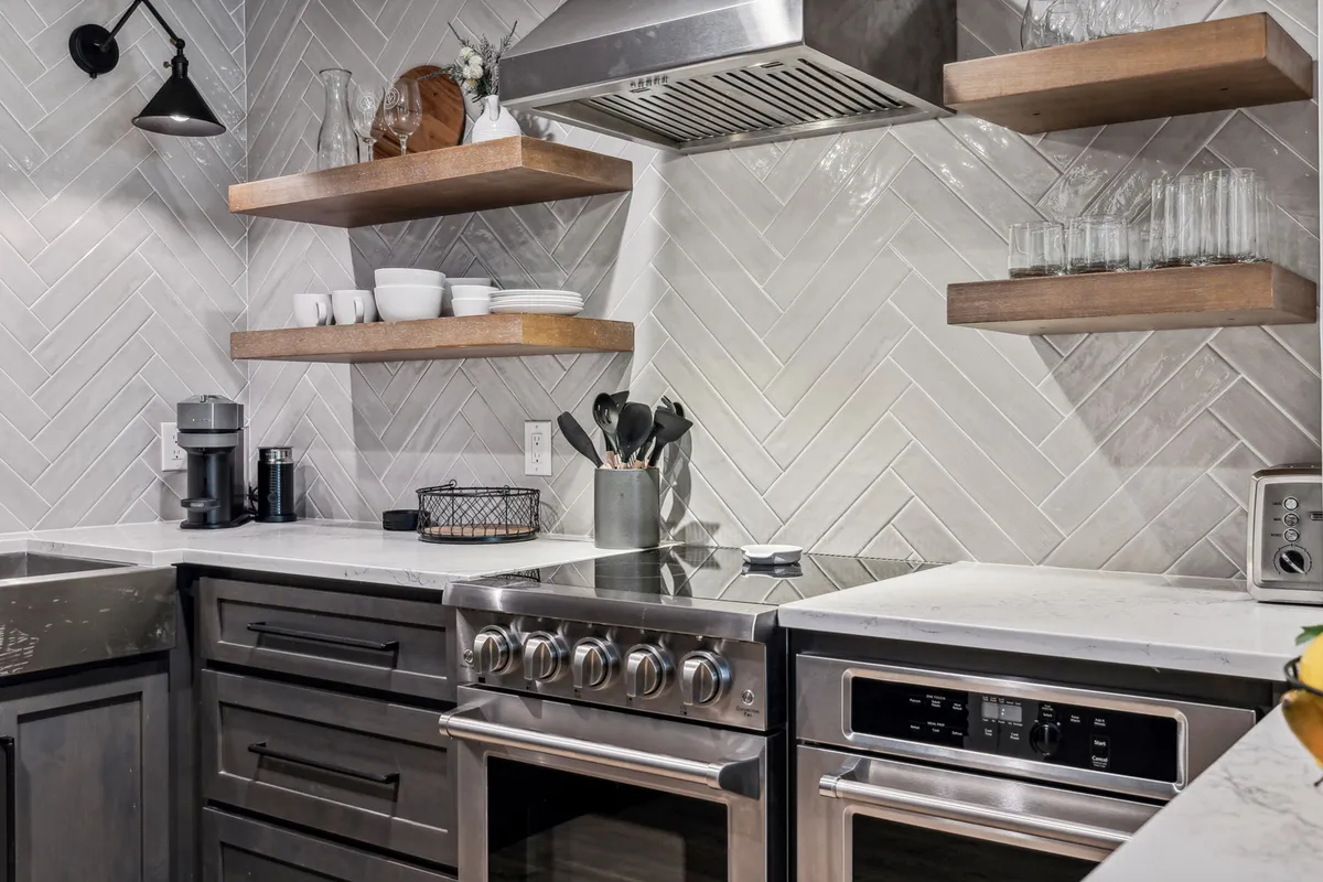 A contemporary kitchen with stainless steel appliances, wooden shelving, a herringbone tile backsplash, and accessible kitchen utensils. The area feels modern, functional, and family-ready.