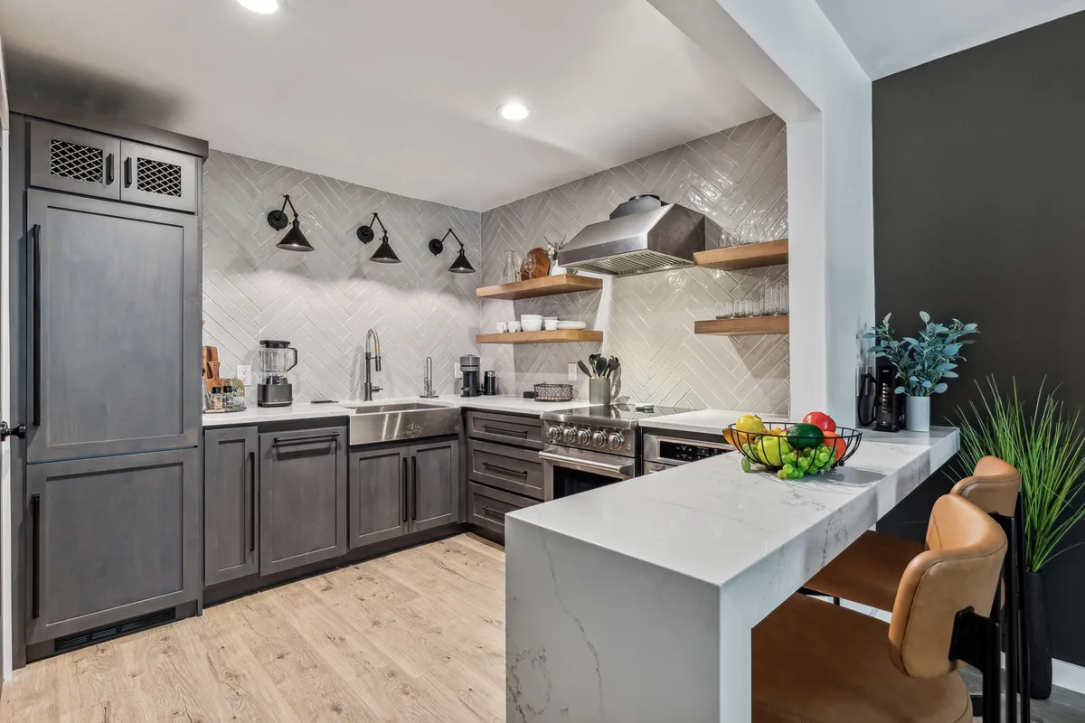 A sleek, modern kitchen featuring stainless steel appliances, a stylish herringbone tile backsplash, marble countertops, and a breakfast bar with comfortable seating. The scene is well-lit and inviting, showing all the essential kitchen amenities ready for meal prep or casual dining.