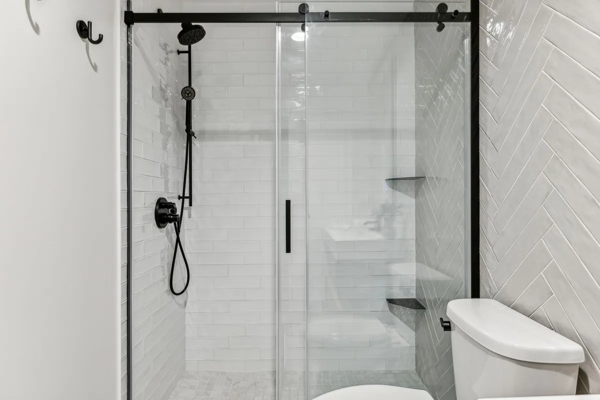 A modern, serene bathroom with a glass shower enclosure, herringbone tile pattern, black fixtures, and a white toilet. The space is fresh, minimalist, and stocked with plush towels, evoking a hotel-like ambiance perfect for a refreshing start or end to your day.
