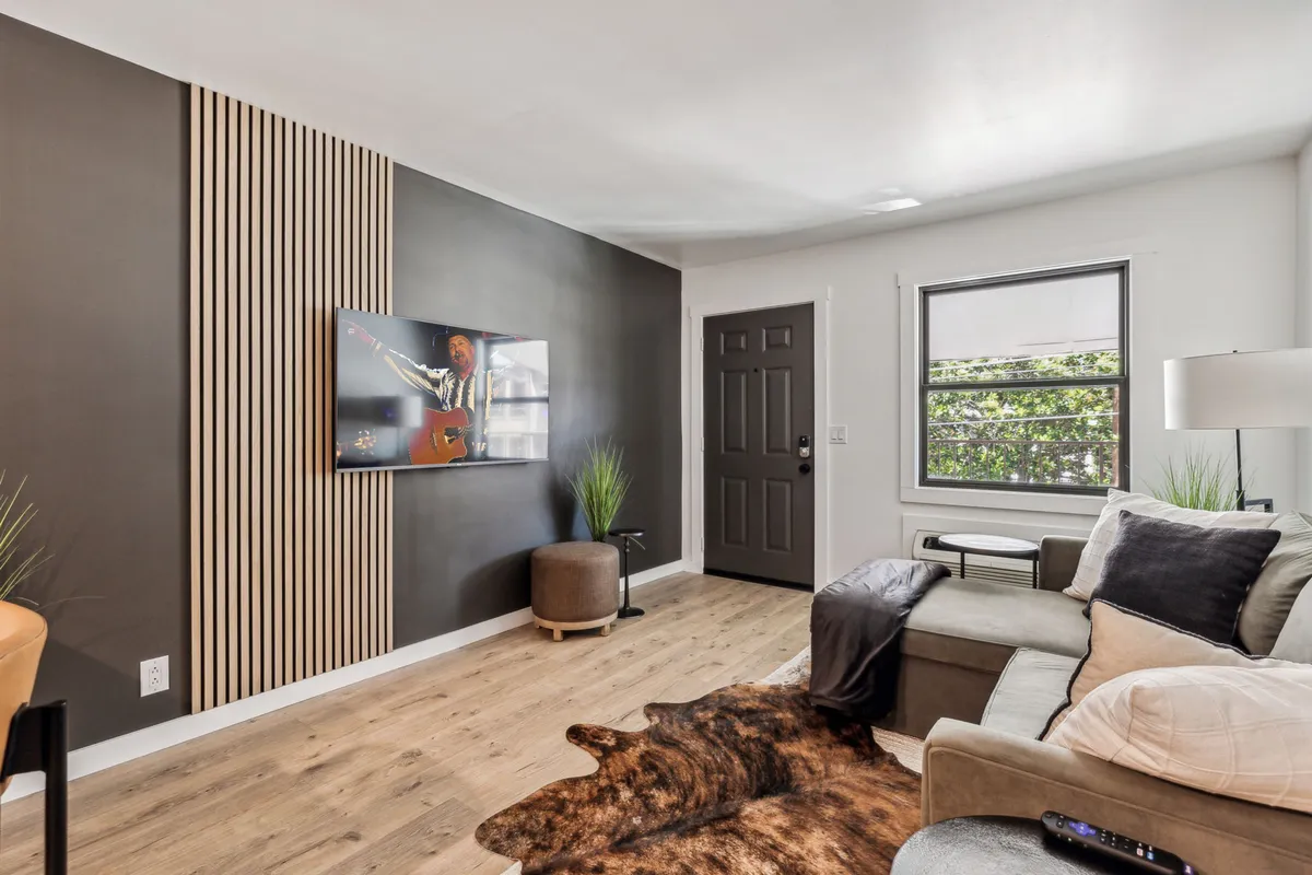 A cozy modern living room featuring a stylish couch with plush cushions, a TV mounted on a wooden slat feature wall, and decorative plants that add a touch of greenery. Warm lighting creates an inviting ambiance, and the space is arranged for comfort and socializing.