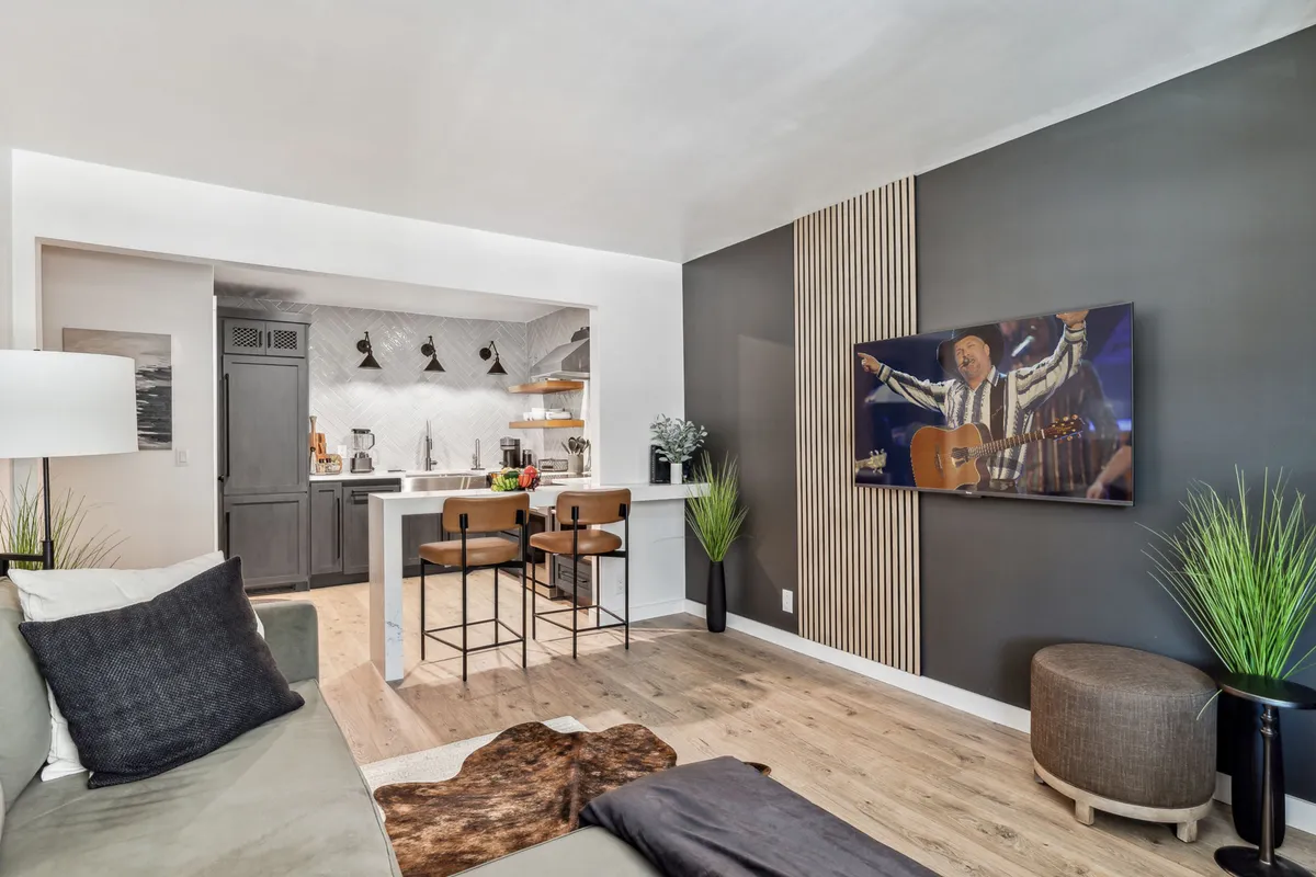 A modern, open-concept living area featuring a plush sofa, sleek kitchen island with bar stools, stylish décor, and inviting greenery. The setup suggests comfort and versatility—perfect for relaxing, entertaining, and enjoying cozy nights in.