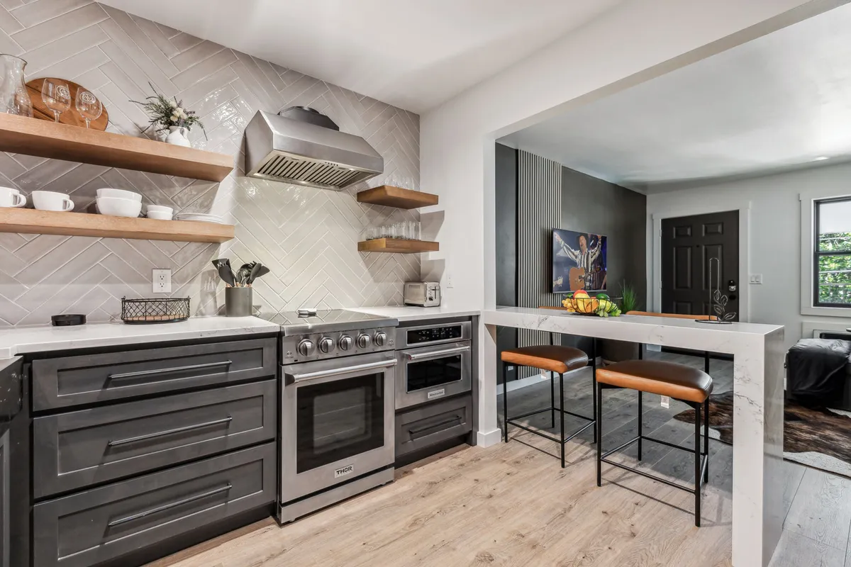 A spacious, contemporary kitchen and dining area featuring modern appliances, open shelving with dishes, elegant bar stools, and a marble countertop. The space is designed for both cooking and socializing, making it versatile for any culinary occasion.