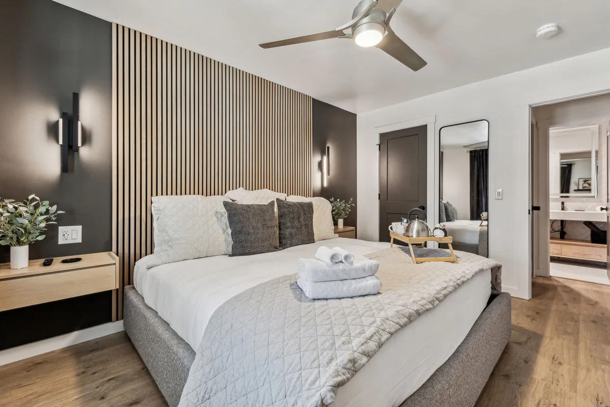 A cozy primary bedroom featuring a king bed with decorative pillows, a warm wooden accent wall, modern lighting fixtures, and a mirror reflecting the spaciousness of the room. Sunlight gently filters through the window, hinting at a relaxing, restful environment ideal for winding down or starting the day.