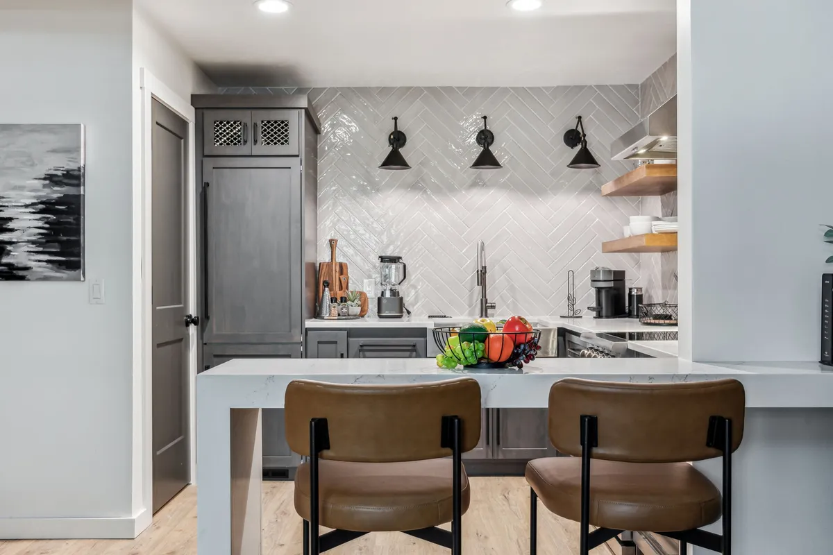 A warm and friendly kitchen scene with a central island, herringbone backsplash, bar stools, and a fruit bowl, making it perfect for socializing or meal preparation. The layout encourages togetherness, with visible amenities that support in-home dining experiences.